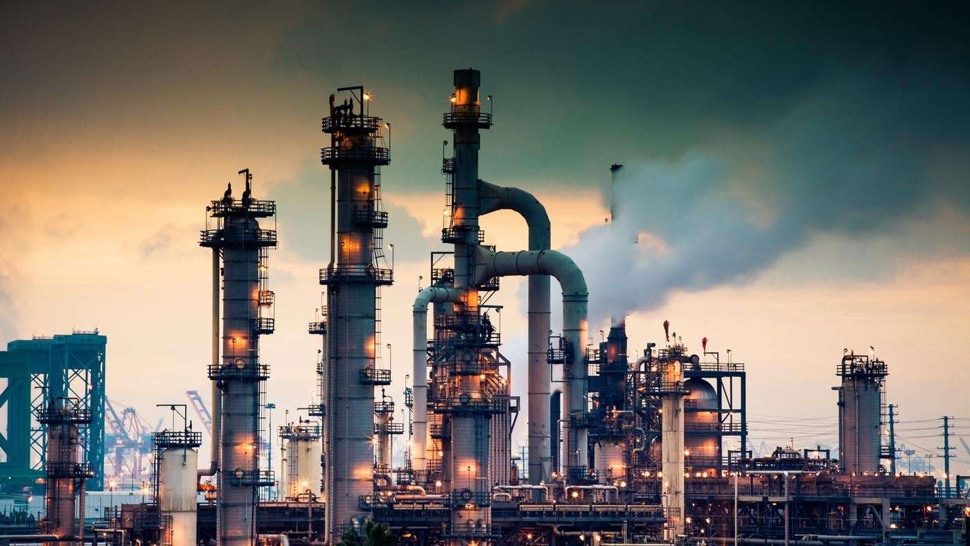 A steaming, glowing mass of smoke stacks and distillation towers connected by conduit and pipe racks towers in the early morning light in front of the Port of Los Angeles.