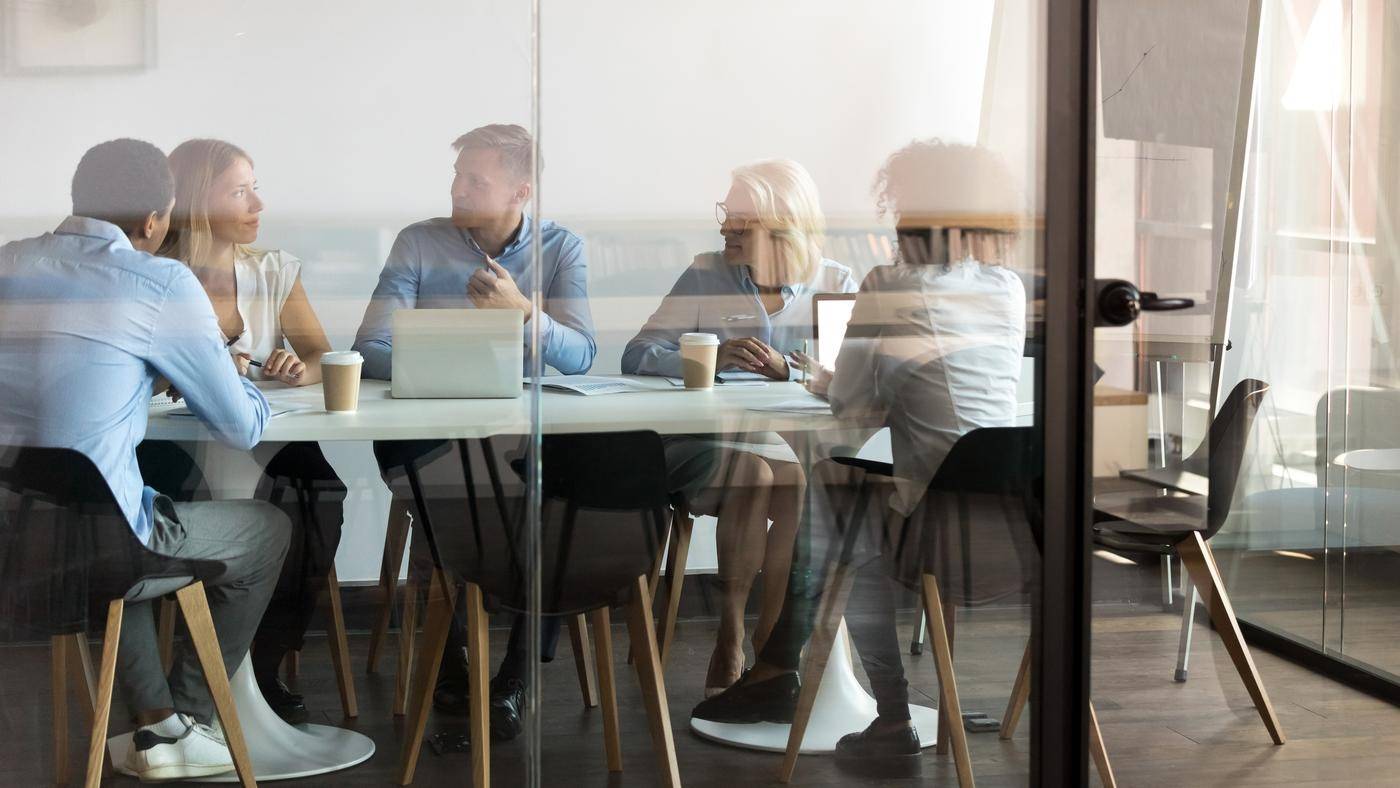 Advertising department brainstorming at modern office boardroom behind closed doors, view through the glass wall. Diverse staff led by ceo discussing new project sharing ideas thoughts and sales pitch