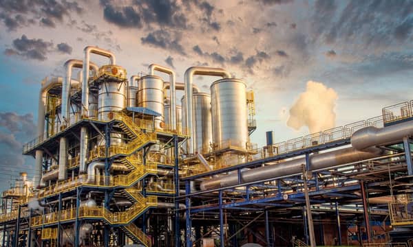 The exterior of a large industrial plant with blue and yellow railings. External stairwells, multiple pipes and large cylinders.