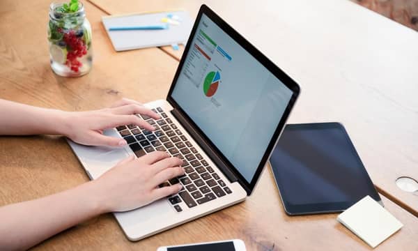 A person checking data on a laptop on a wooden table. iPad and smart phone on the desk.