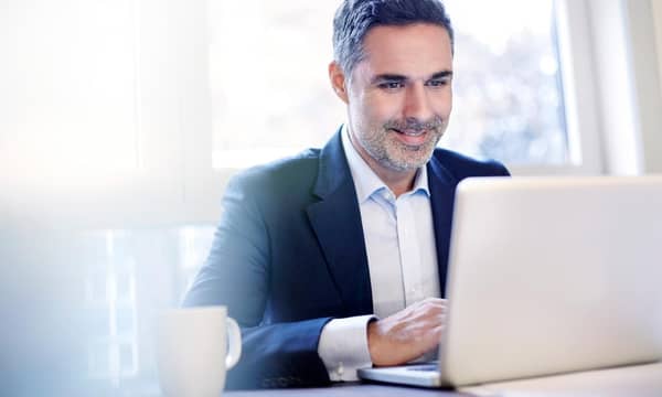 A smiling businessman working on a laptop, in front of window.