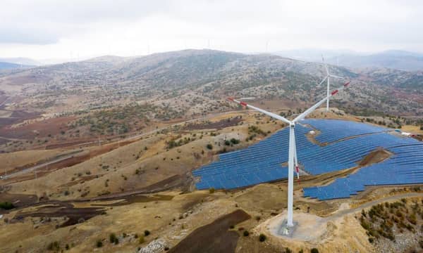 An aerial view of wind turbines and solar panels, across rolling hills.