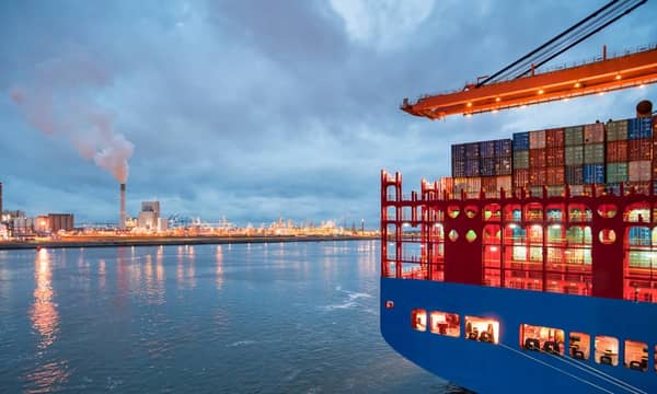 A large container ship docked at a port, next to a large oil refinery.