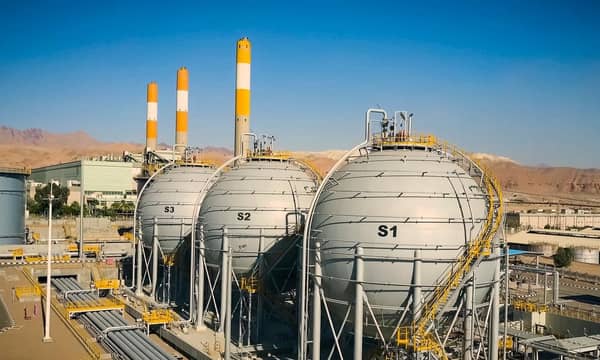 A large power plant with three LNG tanks, mountain range and blue skies in the background.