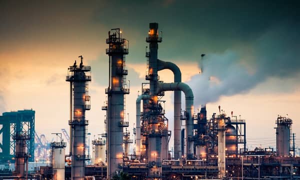 A steaming, glowing mass of smoke stacks and distillation towers connected by conduit and pipe racks towers in the early morning light in front of the Port of Los Angeles.