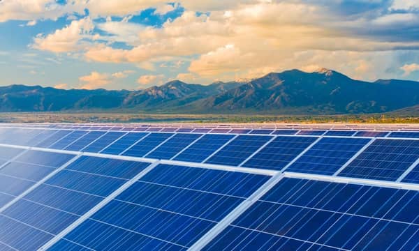 Rows of solar panels in a field, with mountains and blue skies in the background.