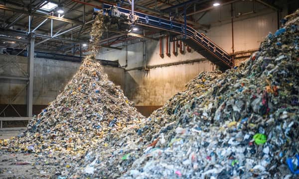 The interior of a concrete warehouse, with an overhanging industrial shredder, processing large amounts of recyclable waste into two vast piles.