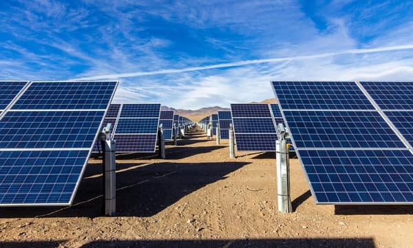 Rows of solar panels in the desert, with a mountainous backdrop and blue skies.