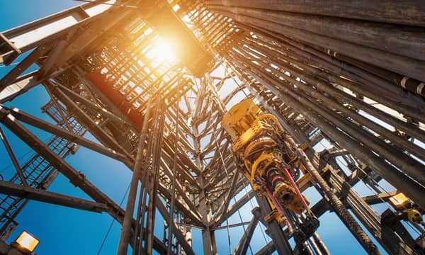An upward view of a central drilling column of an oil rig, the sun and blue skies in the background.