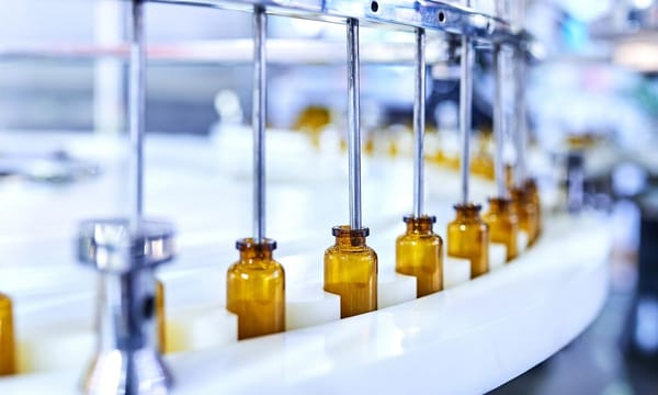 A line of brown glass bottles, on a conveyor belt with metal tubes dispensing a liquid, inside a factory.