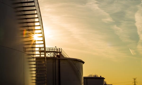 The sun is setting behind a row of oil tanks.