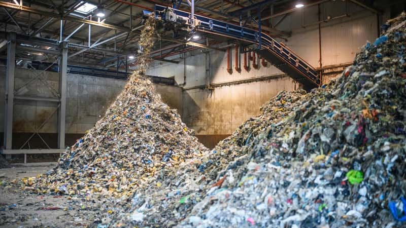 The interior of a concrete warehouse, with an overhanging industrial shredder, processing large amounts of recyclable waste into two vast piles.