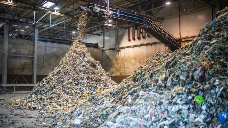 The interior of a concrete warehouse, with an overhanging industrial shredder, processing large amounts of recyclable waste into two vast piles.