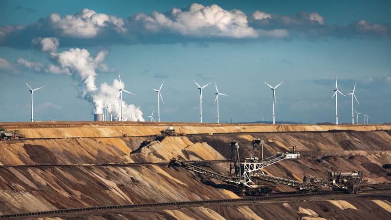 Wind turbines sit atop a quarry, were machines are excavating, with smoke coming from the chimneys of a refinery in the background. 