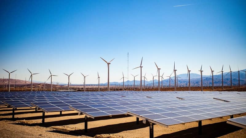 Solar panels and wind turbines in the desert on a backdrop of mountains and clear blue skies.