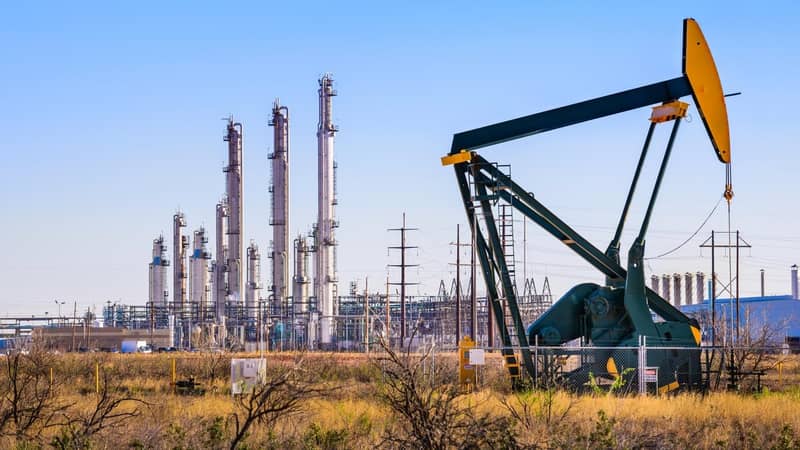 An oil pump in front of an oil refinery, with a network of electricity pylons.