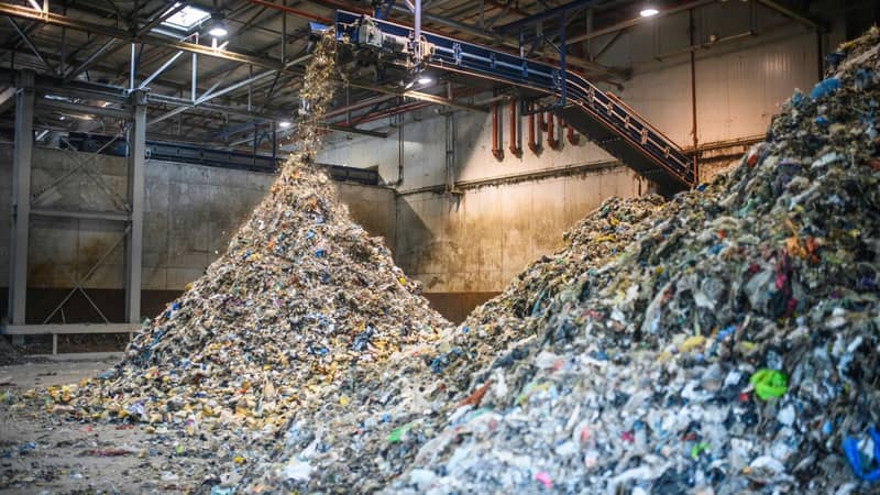 The interior of a concrete warehouse, with an overhanging industrial shredder, processing large amounts of recyclable waste into two vast piles.