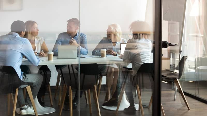 Advertising department brainstorming at modern office boardroom behind closed doors, view through the glass wall. Diverse staff led by ceo discussing new project sharing ideas thoughts and sales pitch