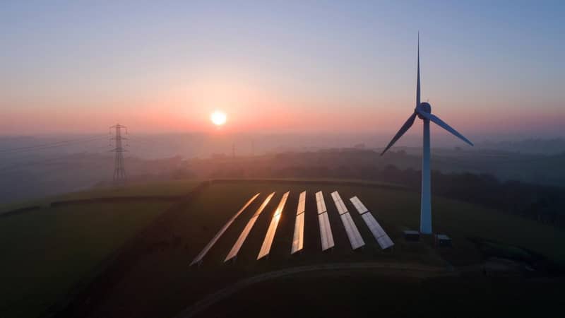 An aerial view of solar panels and a wind turbine in a field at sunset.