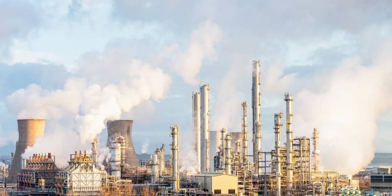 Refinery with chimneys and coal stacks, smoke bellowing out, hazy sky backdrop.