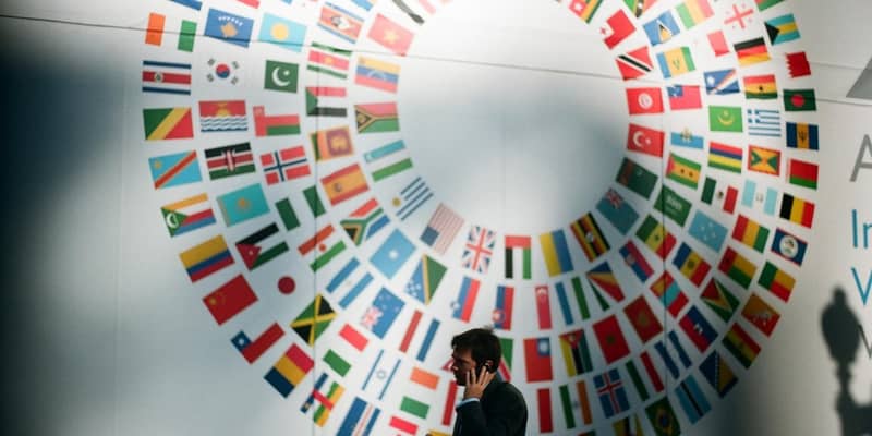 Man on this mobile phone inside a conference room, with a flags of member countries part of the UN on the wall behind.