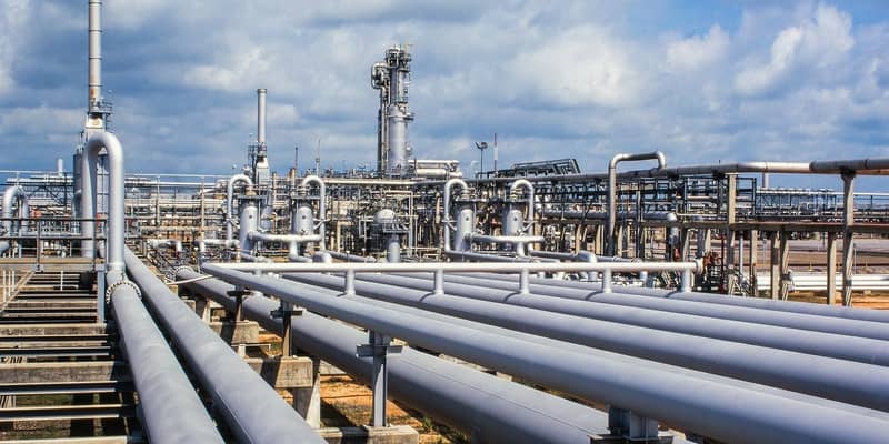Close-up of a gas refinery, a network of pipelines and chimney stacks, against a cloudy blue sky.