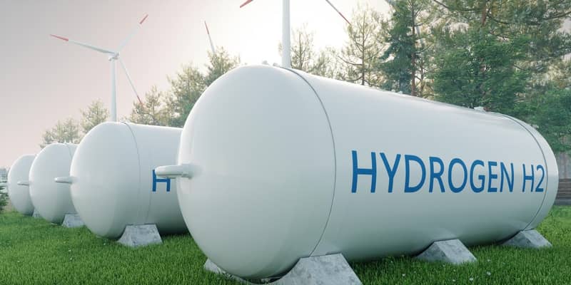 3D render of four hydrogen fuels tanks, in a tree-lined field with wind turbines to the side.