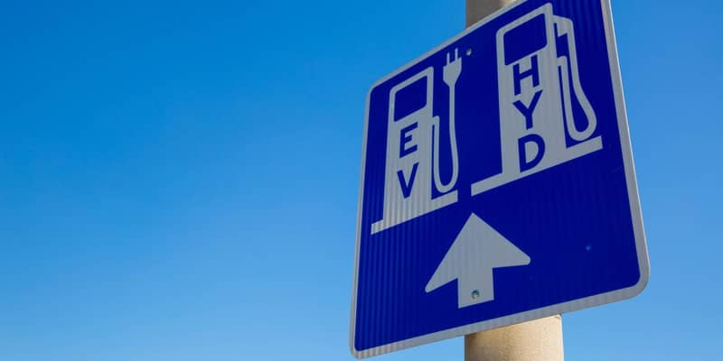 A blue and white road sign on a pole, displaying an area for EV and Hydrogen vehicle charging station.
