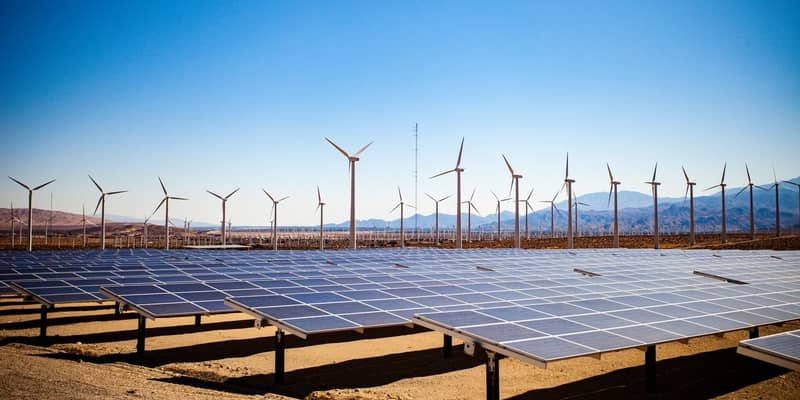 Solar panels and wind turbines in the desert on a backdrop of mountains and clear blue skies.