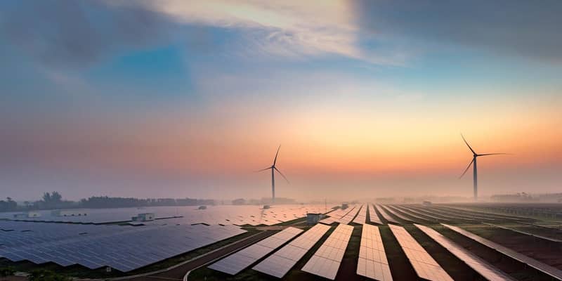 Solar panels and wind turbines across country fields at sunset.