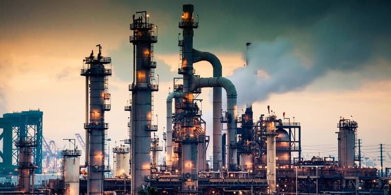 A steaming, glowing mass of smoke stacks and distillation towers connected by conduit and pipe racks towers in the early morning light in front of the Port of Los Angeles.