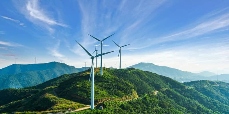 Wind turbines on top of tree covered mountains, against blue skies.