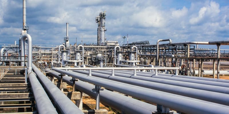 Close-up of a gas refinery, a network of pipelines and chimney stacks, against a cloudy blue sky.