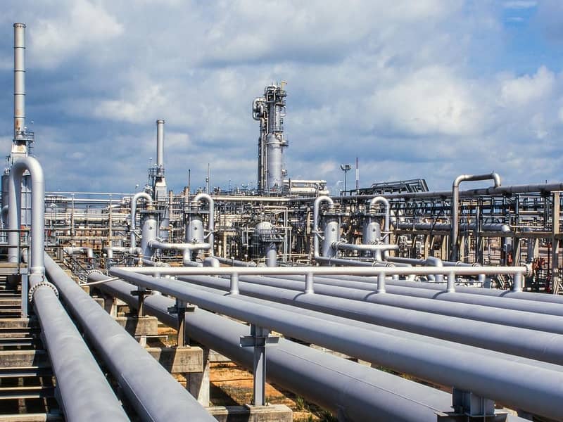 Close-up of a gas refinery, a network of pipelines and chimney stacks, against a cloudy blue sky.