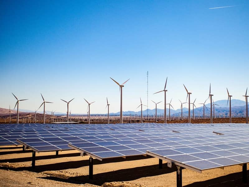 Solar panels and wind turbines in the desert on a backdrop of mountains and clear blue skies.