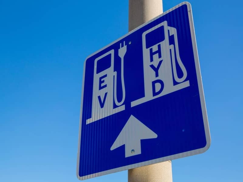 A blue and white road sign on a pole, displaying an area for EV and Hydrogen vehicle charging station.