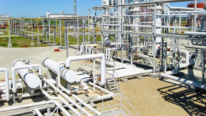 An exterior close-up of an oil refinery with pipes and valves criss-crossing, with a large building and blue skies in the background.