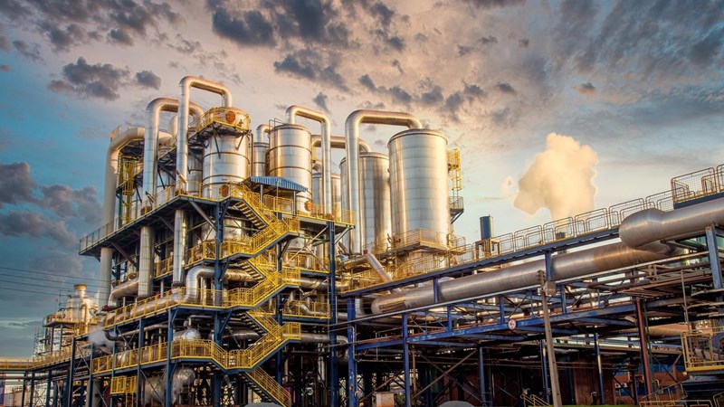 The exterior of a large industrial plant with blue and yellow railings. External stairwells, multiple pipes and large cylinders.