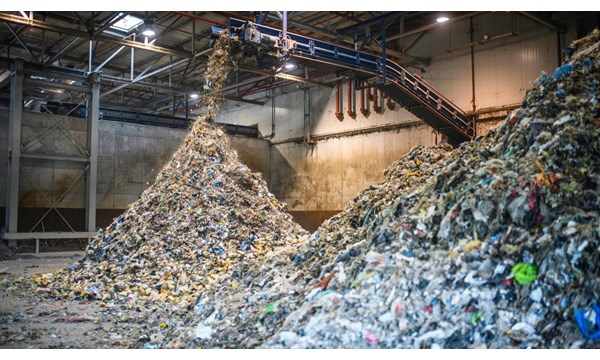 The interior of a concrete warehouse, with an overhanging industrial shredder, processing large amounts of recyclable waste into two vast piles.