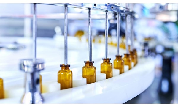 A line of brown glass bottles, on a conveyor belt with metal tubes dispensing a liquid, inside a factory.
