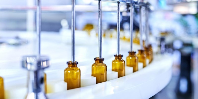 A line of brown glass bottles, on a conveyor belt with metal tubes dispensing a liquid, inside a factory.
