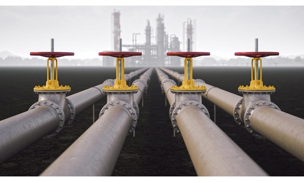 A set of 4 large pipes with red valves, outside and leading to an industrial plant in the background.