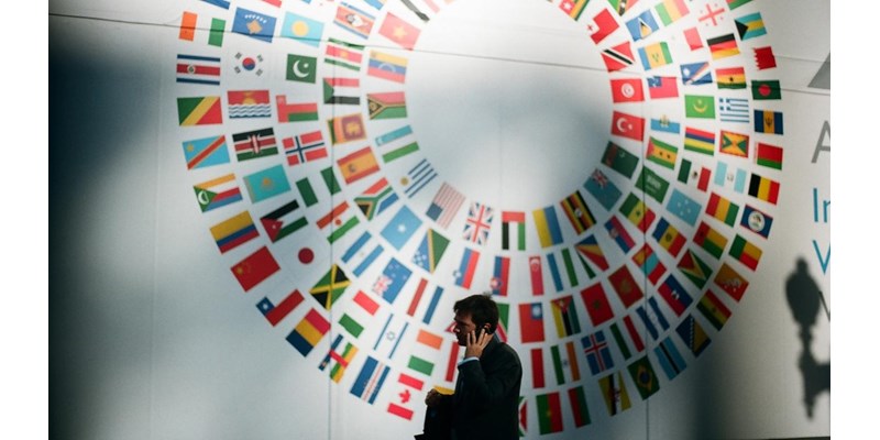 Man on this mobile phone inside a conference room, with a flags of member countries part of the UN on the wall behind.