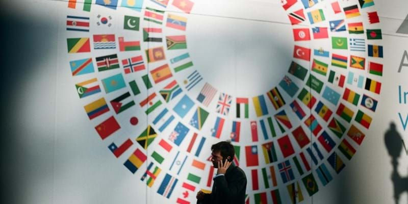 Man on this mobile phone inside a conference room, with a flags of member countries part of the UN on the wall behind.