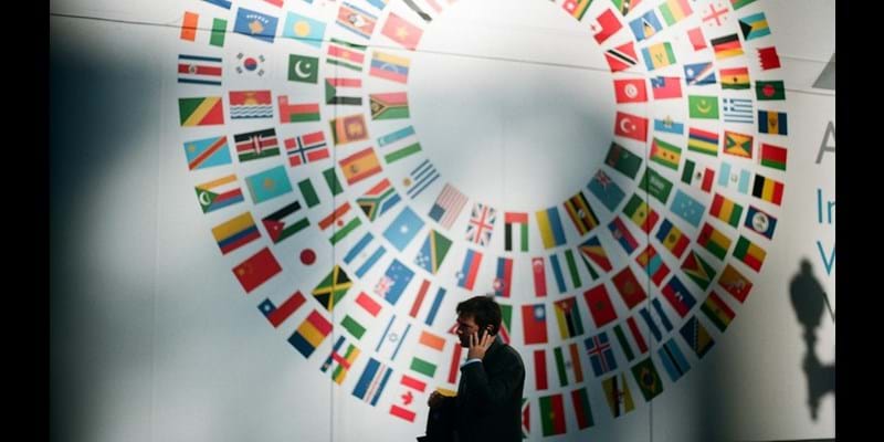 Man on this mobile phone inside a conference room, with a flags of member countries part of the UN on the wall behind.