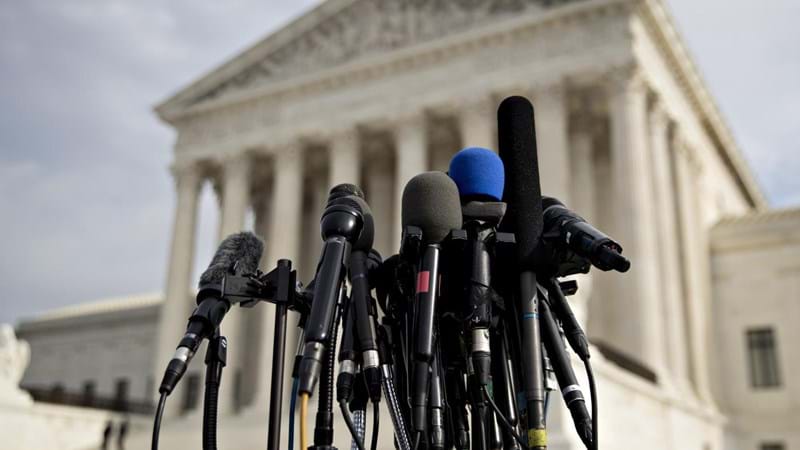 Close-up of a bank of media microphones, outside a neoclassical building.