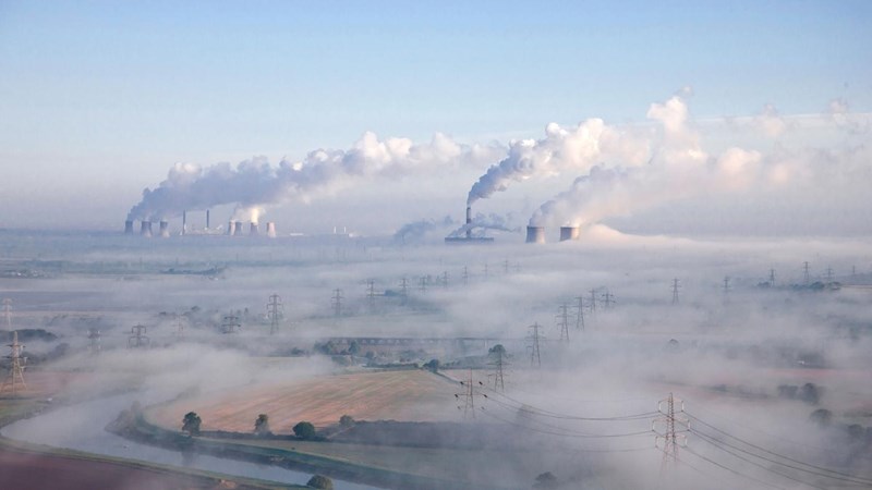 A sprawling refinery, blanketed in smoke from the industrial chimney stacks. Country fields, stream and electricity pylons in the foreground.
