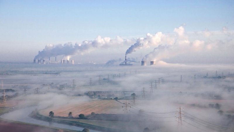 A sprawling refinery, blanketed in smoke from the industrial chimney stacks. Country fields, stream and electricity pylons in the foreground.