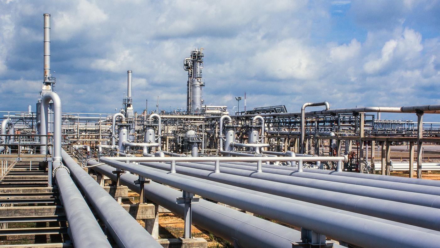 Close-up of a gas refinery, a network of pipelines and chimney stacks, against a cloudy blue sky.