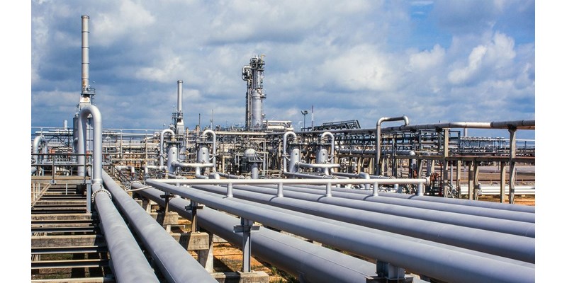 Close-up of a gas refinery, a network of pipelines and chimney stacks, against a cloudy blue sky.